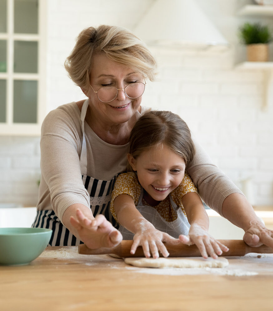 grandmother rolling out dough with her granddaughter covered in flour